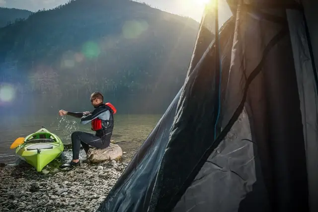 A paddler resting on a rock beside a green kayak on a mountain lake shore, a tent in the foreground